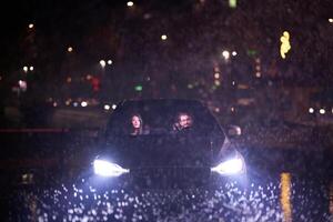 In the midst of a nighttime journey, a happy family enjoys playful moments inside a car as they travel through rainy weather, illuminated by the glow of headlights, laughter, and bonding photo