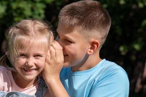 Sibling secrets unfold in a grassy glade, as a boy leans in to share a whisper with his grinning sister, used for National siblings day, happiness in nature and escapism the,es. photo
