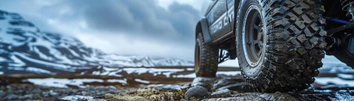 Rugged offroad vehicle tire on a rocky terrain, blurred snowy mountain background, dramatic overcast sky photo