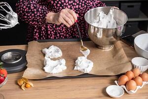 A baker in a floral shirt expertly sculpts peaks of meringue on a baking sheet, ideal for illustrating pastry technique in cooking guides. photo