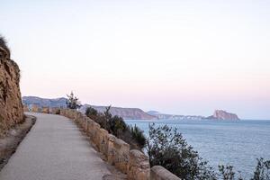 A coastal pathway flanked by a stone balustrade leads towards a soft-hued sunset over distant cliffs and a calm sea. photo