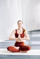 A slender woman in a brown sports uniform sits in the lotus position against a background of white curtains and meditates, front view. Yoga classes in the loft studio photo