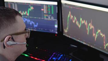 Exchange stock market. Back view of young professional trader in eyeglasses pointing on the data on computer screen with pen and holding Bitcoin in one hand while working his modern office video