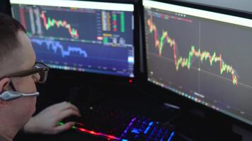 Exchange stock market. Back view of young professional trader in eyeglasses pointing on the data on computer screen with pen and holding Bitcoin in one hand while working his modern office video