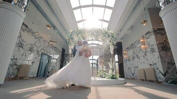 The bride and groom are dancing under a flower arch. The groom is in a black tuxedo, and the bride in white gown with a veil. The backdrop features marble walls and modern lighting. video