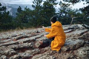 A person in a yellow raincoat sitting on top of a mountain with hands raised in awe of nature's photo