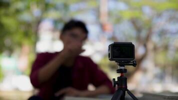 Asian man recording and talking to camera. Young man vlogging and taking selfie with action camera at public park. Action camera recording a man with blurry background video