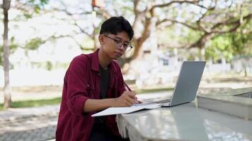 asiatisch Hochschule Schüler studieren auf Laptop beim Campus draussen Park. Mann Schreiben auf ein Hinweis Buch und Arbeiten auf Laptop. lehrreich Konzept video