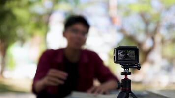 Asian man recording and talking to camera. Young man vlogging and taking selfie with action camera at public park. Action camera recording a man with blurry background video