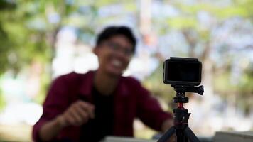 Asian man recording and talking to camera. Young man vlogging and taking selfie with action camera at public park. Action camera recording a man with blurry background video