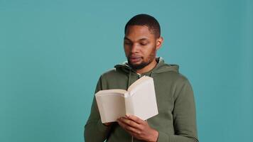 Man laughing while reading funny book, entertained by engrossing story. Joyful person having fun, being amused by literature novel during leisure time in studio background, camera A video