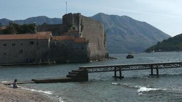 Montenegro, Budva - July 29, 2021. Shore with waves on background of medieval fortress. Creative. Beautiful beach with tourists and fortress with mountains on horizon. Medieval citadel on shore with video