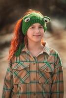 portrait of a cute red-haired teenage girl with freckles wearing a green knitted hat in the form of a frog photo