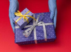 Hands in blue latex gloves holds gift boxes in color paper with polka dots with tied ribbon with as a present for Christmas, new year, valentine day or anniversary on red background. Top view photo