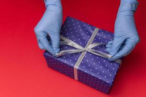 Hands in blue latex gloves holds gift box in color paper with polka dots with tied ribbon with as a present for Christmas, new year, valentine day or anniversary on red background. Top view photo