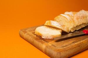 Sliced bread baguette on a wooden cutting board with knife on a terracote color background. Copy space. Close-up high quality photo