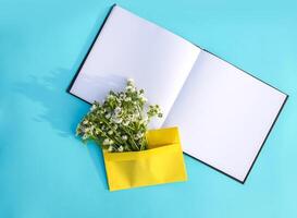 Yellow paper envelope with small garden white chamomile flowers and empty notepad on light blue background. Festive floral template. Greeting card design. Top view. photo