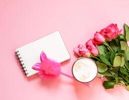 The cup of hot coffee, empty notepad with funny pen and bouquet of fresh roses on soft pink background. photo