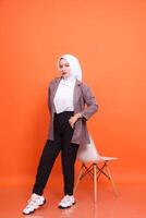 Full length portrait of adult Muslim indonesia woman standing with her back to chair with arms crossed, wearing suit office clothes isolated on orange studio background. Concept of fashion, company photo