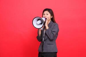 Asian office woman's expression tilted to the right, shouting angrily using a megaphone, hand focusing her voice, wearing a gray jacket and skirt on a red background. for broadcast and advertising photo