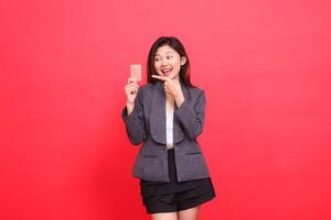 Cheerful candid expression of Asian office girl in hand holding debit credit card while pointing at it wearing jacket and skirt on red background. for financial, business and advertising concepts photo
