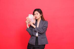 The expression of a cheerful Asian girl director with both hands holding a credit card, debit card and money kissing the camera wearing a gray jacket and skirt on a red background. for advertising photo