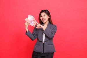 Asian office woman smiling expression holding credit, debit card and money while pointing top right at the camera wearing gray jacket and red skirt. for transaction, business and advertising concepts photo