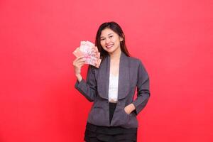 Expression of cheerful indonesia office woman holding debit credit card and money upright and in pocket looking at camera wearing gray jacket and red skirt. for transaction, business and advertising photo