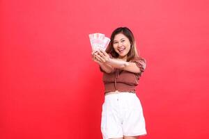 gesture of a cheerful young Asian woman with both hands holding rupiah money in front of her wearing a brown blouse with a red background. for transaction, technology and advertising concepts photo