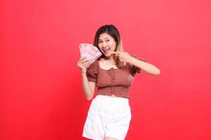the expression of a cheerful, happy Asian woman holding rupiah money while pointing at it, wearing a brown blouse on a red background. for transaction, technology and advertising concepts photo