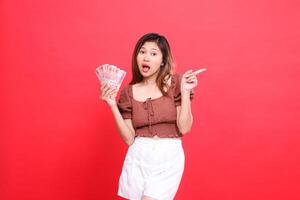 The expression of a shocked Asian woman holding rupiah money while pointing to the left wearing a brown blouse with a red background. for transaction, technology and advertising concepts photo