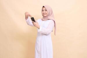 woman asian muslim wearing a hijab smiles cheerfully at the camera, operating a cellphone gadget with both hands while pointing. Lifestyle, technology and promotion concept photo