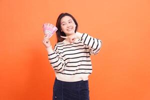 an indonesia woman with a candid cheerful expression standing while holding a number of rupiah bills while pointing at them. for fashion, business and finance concept photo