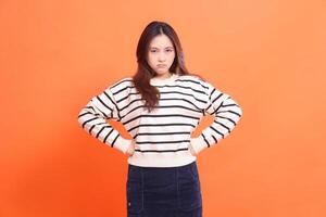 beautiful indonesia girl pouting annoyed confident standing with hands clenched to waist and looking at camera isolated on orange background wearing white sweater photo
