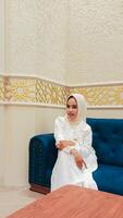 beautiful asian woman happy expression both hands crossed and resting on thighs and sitting on sofa with islamic ornament calligraphy background arabic atmosphere photo
