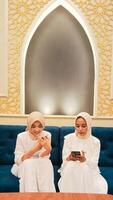 a pair of women sitting on a sofa operating their respective cellphone gadgets with an elegant Arabic Muslim calligraphy background photo