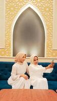 portrait of a pair of women sitting on a sofa smiling selfie in front of a cellphone camera with an elegant Arabic Islamic calligraphy background photo