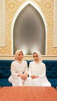 portrait of a pair of women sitting on a sofa smiling gracefully with a welcome hand gesture sawadikap facing the camera with an elegant Arabic Islamic calligraphy background photo