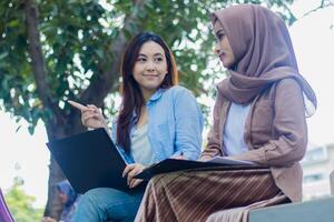 a pair of graceful indonesia women candidly show something the part left when discussing the task of carrying a laptop, books and clipboard in the campus park. students for education concepts photo