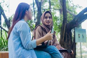 two candid Asian women smiling discussing studying together holding laptop, books and clipboard in campus park. female students for education, promotion and lifestyle concepts photo