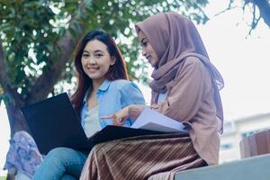 A pair of beautiful Asian girl looking at the camera are discussing together carrying laptops, books and clipboards while pointing in the campus park. students for education and lifestyle concepts photo