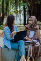 A pair of indonesia women candidly discussing school assignments carrying laptops, books and clipboards in the campus park. female students for education, promotion and lifestyle concepts photo