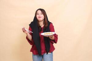 beautiful mature Asian woman wearing a red hijab, smiling, eating dimsum using chopsticks and carrying a plate containing dimsum Chinese food candid. used for food, health and culinary content photo