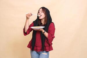 Beautiful mature indonesia woman wearing a red hijab poses, bites into dimsum with chopsticks up and carries a plate containing dimsum Chinese food. used for food, health and culinary content photo
