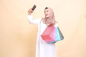 A young Asian Muslim woman wearing a cheerful hijab takes a selfie in front of a cellphone camera and carries a paper bag. for advertising, culture, Eid and Ramadan photo