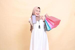 a young Asian Muslim woman in a happy hijab displays a cellphone screen and carries a paperbag shopping bag. for advertising, technology, Eid and Ramadan photo