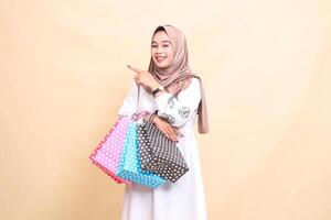 the story of a young Asian Muslim woman wearing a cheerful hijab pointing to the right and carrying a paperbag shopping bag. for advertising, culture, Eid and Ramadan photo