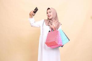 a young indonesia Muslim woman wearing a cheerful hijab on a call greets in front of a cellphone camera and carries a paper bag shopping bag. for advertising, culture, Eid and Ramadan photo