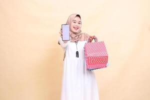 a young Asian Muslim woman in a happy hijab displays a cellphone screen and carries a paperbag shopping bag. for advertising, technology, Eid and Ramadan photo