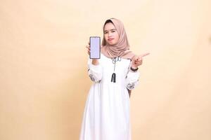 An indonesia Muslim girl wearing a hijab frowns showing a cell phone screen gadget pointing left to create sales content. for advertising, technology, Eid and Ramadan photo
